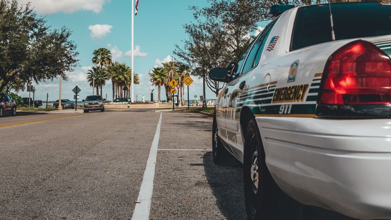 A police car parked by a road under a clear sky with an American flag and trees in the background.