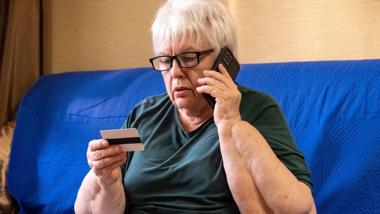 Senior woman in glasses sitting on sofa looking on credit card and talking on phone. Concept of scam, money loss, banking protection, finance fraud, customer service, remote payment