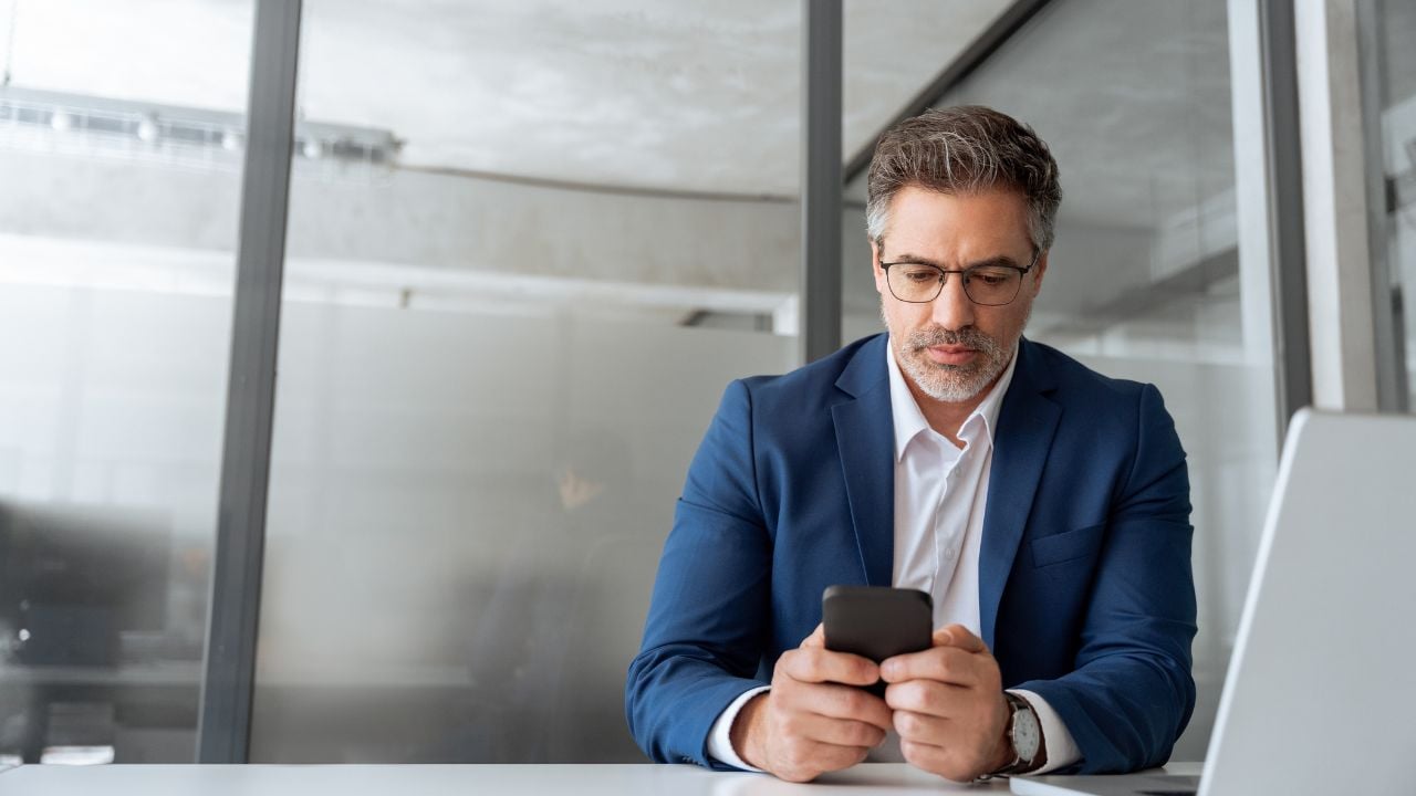 Busy mature ceo businessman in suit using phone for business work in office. Worried senior finance professional man working on cellphone telephone, pc laptop computer looking at screen. Copy space